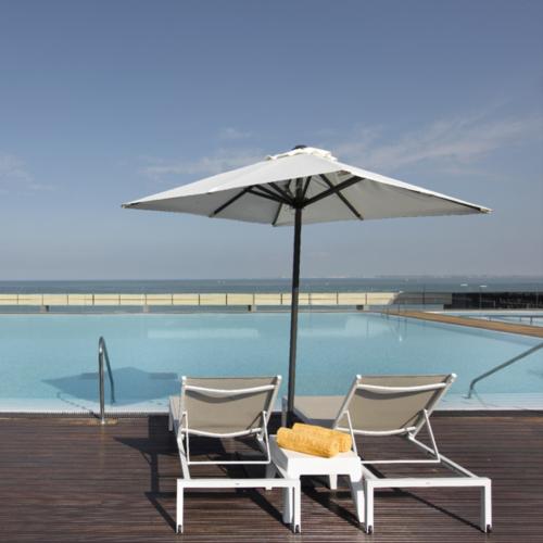 Imagen de la piscina del Parador de Cádiz con dos tumbonas bajo una sombrilla y al frente una gran piscina azul frente al mar